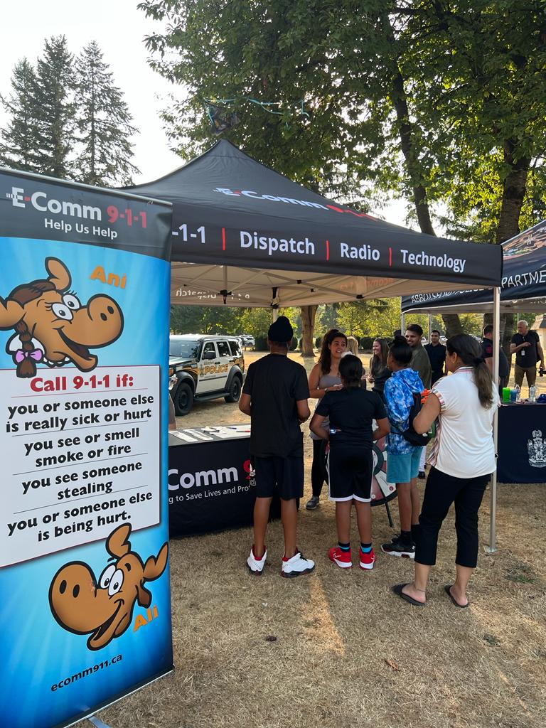 Communications Specialist Martina sharing important 9-1-1 info with visitors at the Abbotsford Police Movie Night on September 2. Several kids and a parent are lined up at the booth to spin E-Comm's trivia wheel and respond to a question about 9-1-1 and earn a 9-1-1 sticker.