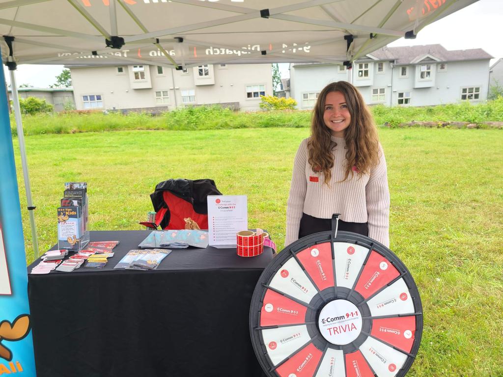 Communications Specialist Kelly Furey attending Hats Off Day in Burnaby on June 18. She is standing behind E-Comm's trivia wheel and presenting our brochures and merchandise.
